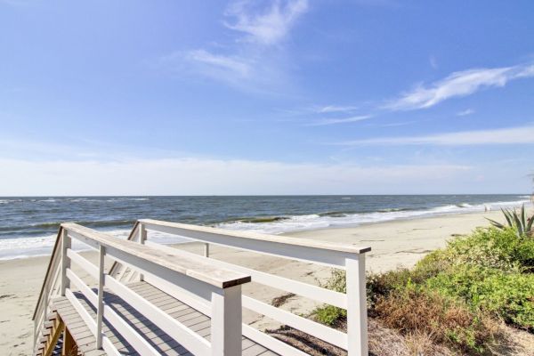 A wooden walkway leads to a sandy beach with gentle waves under a clear blue sky, surrounded by some greenery.