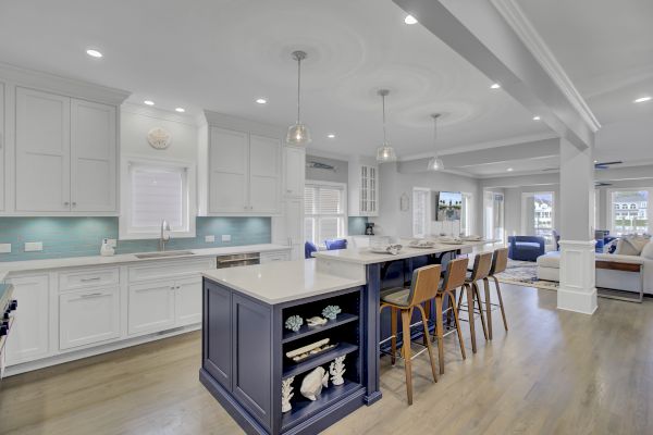A modern kitchen with a large island, bar stools, pendant lights, and white cabinets opening to a living area.