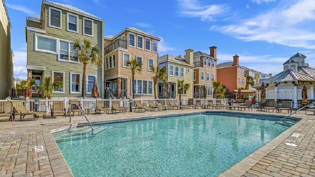A swimming pool surrounded by colorful three-story houses and palm trees under a clear blue sky.