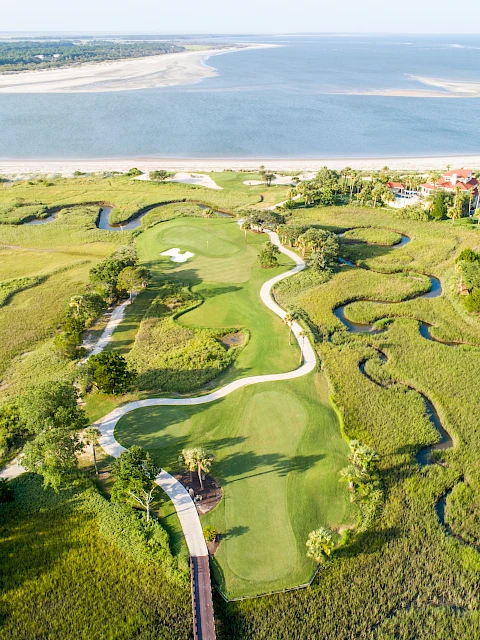 The image shows an aerial view of a coastal golf course with winding paths and surrounding grassy marshlands, near a body of water.