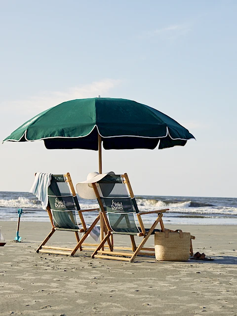 Two beach chairs under a green umbrella facing the ocean, with a straw hat and beach bag nearby on the sand.