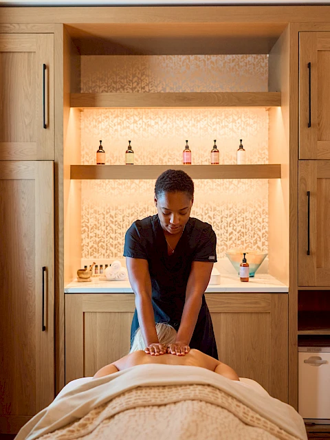 A person giving a massage to someone lying face down on a massage table in a cozy spa room with wooden cabinetry and a small fridge, light streaming in.