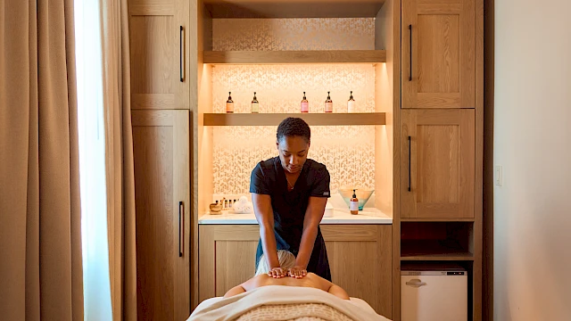 A person giving a massage to someone lying face down on a massage table in a cozy spa room with wooden cabinetry and a small fridge, light streaming in.