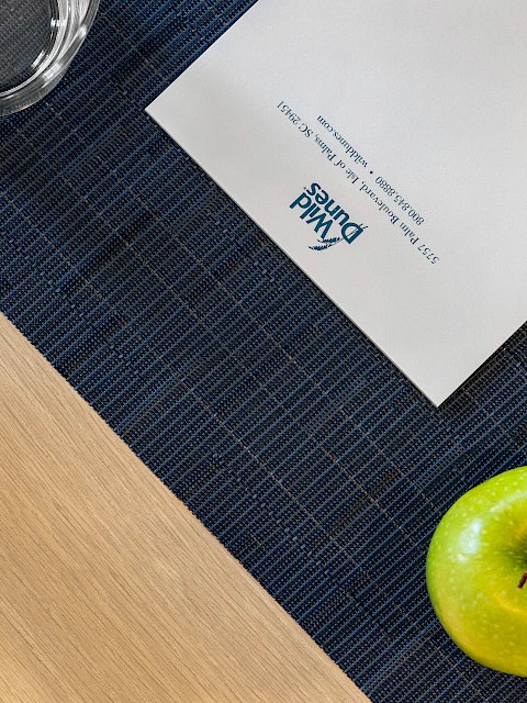 A green apple, pen, glass, paper with "Wild Dunes" logo, and a plant on a wooden desk with a blue mat.