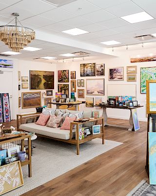 The image shows an art gallery with paintings displayed on walls and easels, and a seating area with couches and tables.