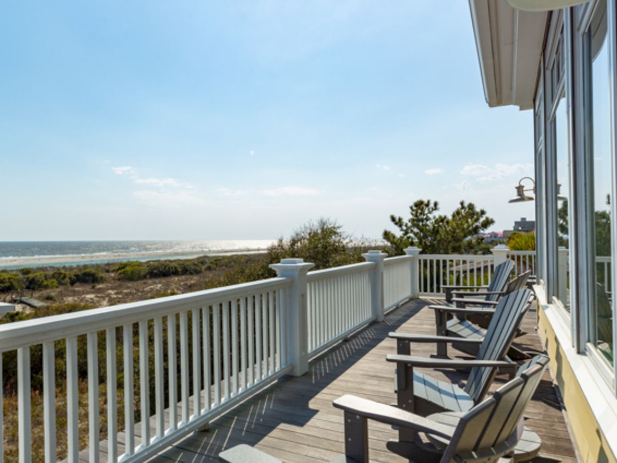 A deck with wooden chairs overlooks the ocean and sandy terrain, under a clear blue sky, next to a building with large windows.