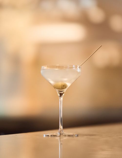 A delicate martini glass sits on a reflective bar, a thin olive skewered by a pick, warm blurred lights creating a classy evening mood.