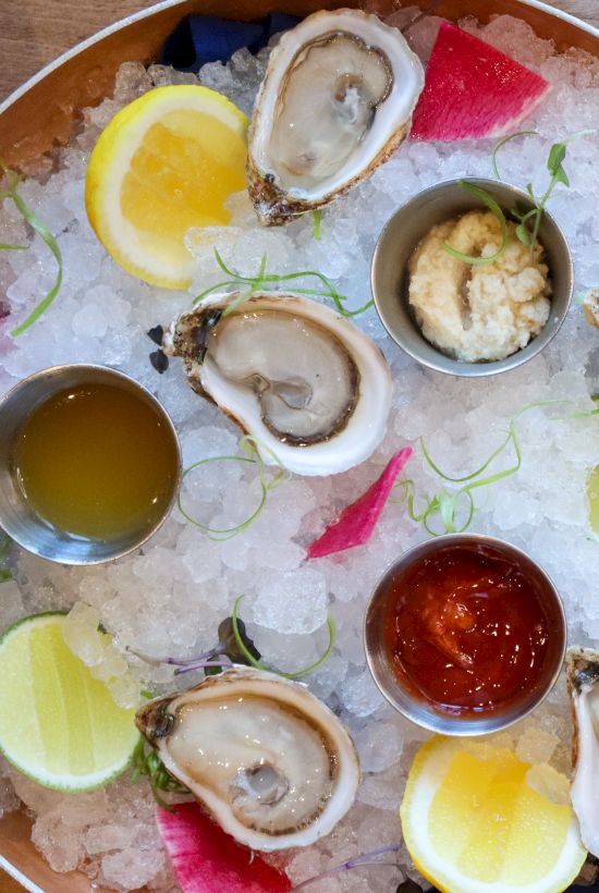 A platter with oysters, lemon, sauces, a plate with bread and cured meat, and another with sliced lime and food on a black slate.