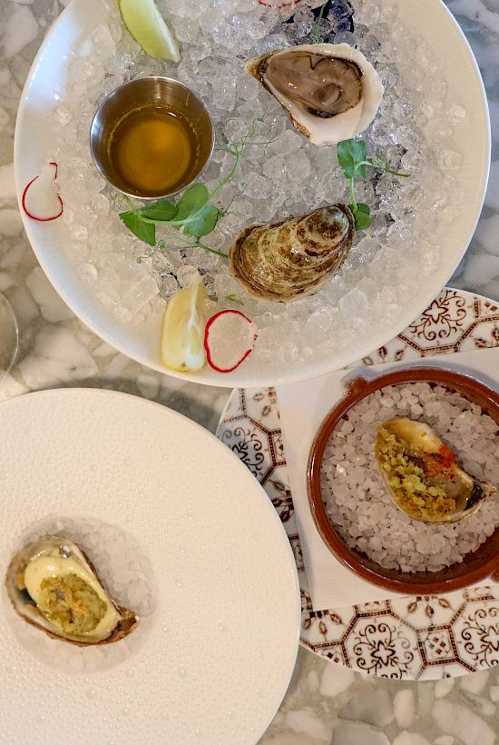 The image shows two plates with oysters, a glass of white wine, and a small bowl with sauce on a marble table.
