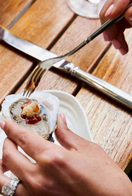 A person holds an oyster with sauce, using a fork to eat, on a wooden table setting.