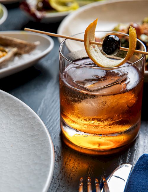 A cocktail garnished with an olive and citrus peel on a skewer is centered, surrounded by plates of food on a table setting.