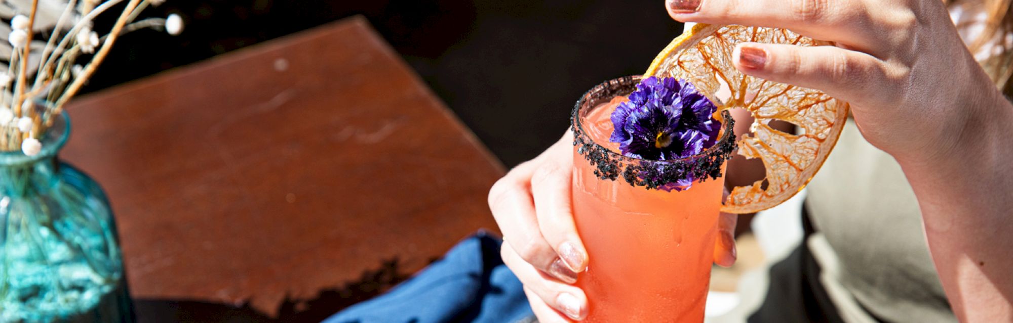 A person holds a pink drink garnished with a dried orange slice and purple flower, next to a teal vase on a table.