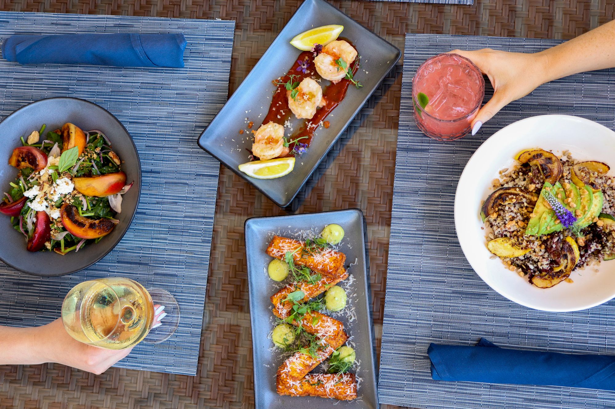 An overhead shot of a table with assorted dishes, including salads and appetizers, and two beverages in hand, set on blue placemats.
