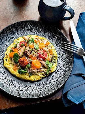 A colorful omelette with vegetables on a plate, accompanied by a cup of coffee, a fork, and a knife on a blue napkin ends the setting.