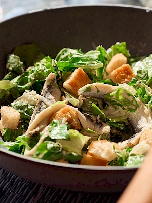 A salad with greens, cucumber, croutons, and shaved cheese in a dark bowl, sunlight on the table, a hand in view, fresh and vibrant.