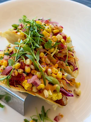 A taco topped with corn, diced onions, greens, and other colorful ingredients is placed on a white plate with a taco holder.