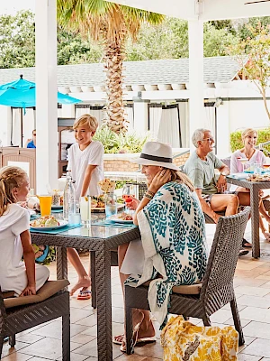 People enjoy a sunny day at an outdoor restaurant, sitting and chatting around tables with food and drinks, surrounded by greenery.