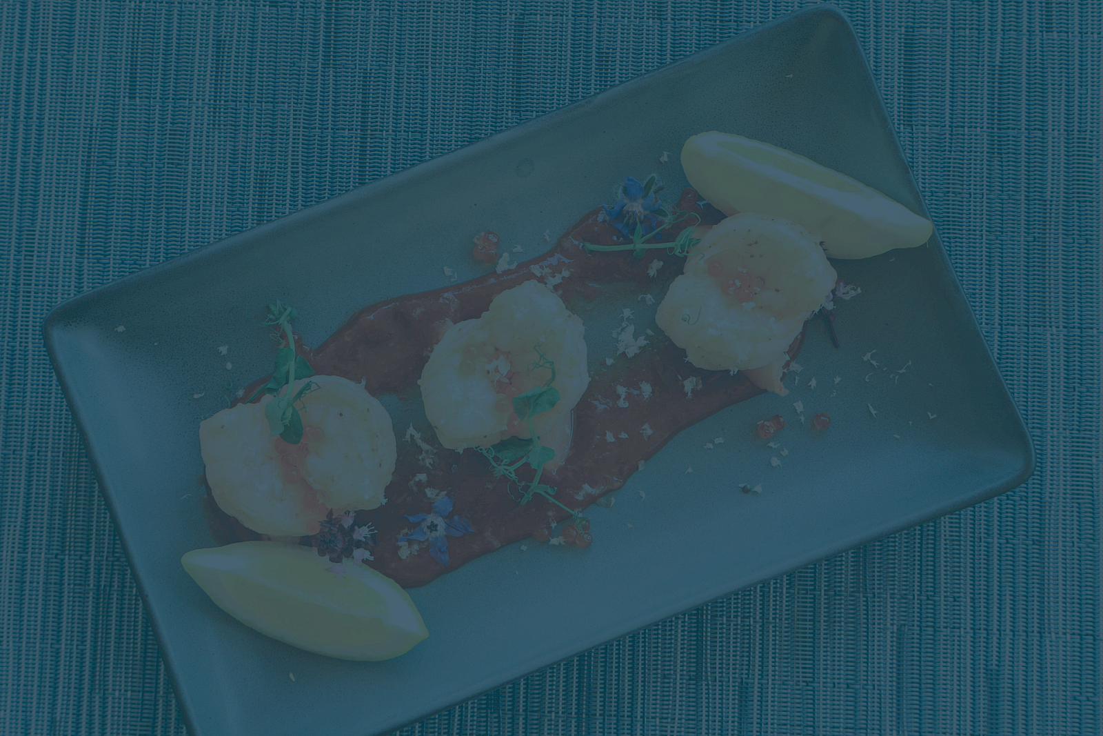 A dish with three shrimp on a red sauce, garnished with herbs, flowers, and lemon wedges, on a rectangular plate.