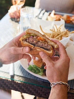 Two people share a grilled sandwich at an outdoor table with fries, salad, and drinks visible, enjoying a casual meal together.