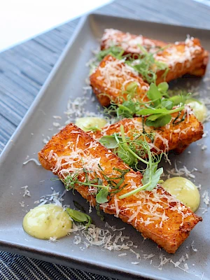 The image shows a plate with crispy fried pieces topped with grated cheese and fresh herbs, accompanied by dollops of yellow sauce.