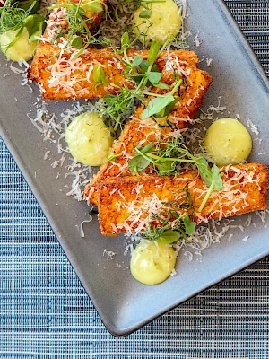 The image shows a dish with roasted carrots, garnished with microgreens, grated cheese, and dollops of a yellow sauce, on a rectangular plate.