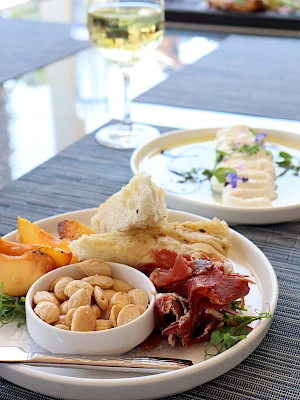 A plate with nuts, cured meat, bread, and cheese, accompanied by a glass of white wine, on a table with another dish in the background.