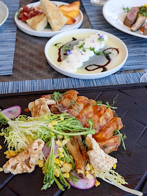 A vibrant restaurant spread: crispy grilled meat with greens on a slate plate, a creamy dip with drizzle, and assorted appetizers including fried items and a colorful salad, all set on a wooden table.