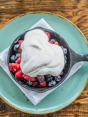 A skillet dessert with mixed berries and a generous dollop of whipped cream sits on a green plate atop a wooden table.