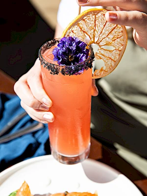 A hand holding a pink cocktail garnished with a dried citrus slice and purple flowers, with a plate of food partially visible below.