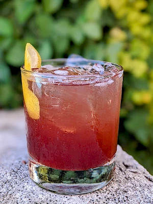 A cocktail with a reddish gradient, ice cubes, and a lemon twist garnish in a glass, set against a blurred green background.