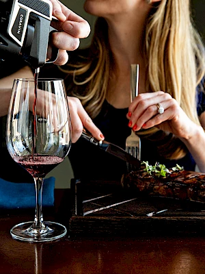 A person cuts a steak while another hand pours wine into a glass, creating a cozy dining atmosphere.
