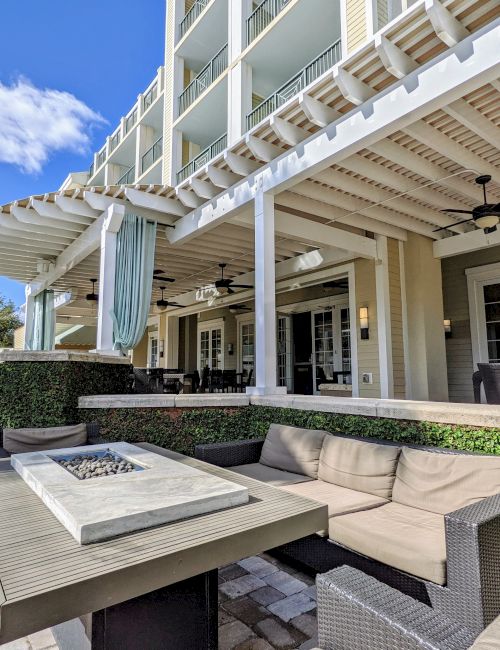 Outdoor terrace with seating, a fire pit, and a palm tree under a blue sky. There's a sign for "The Terrace" on the canopy.
