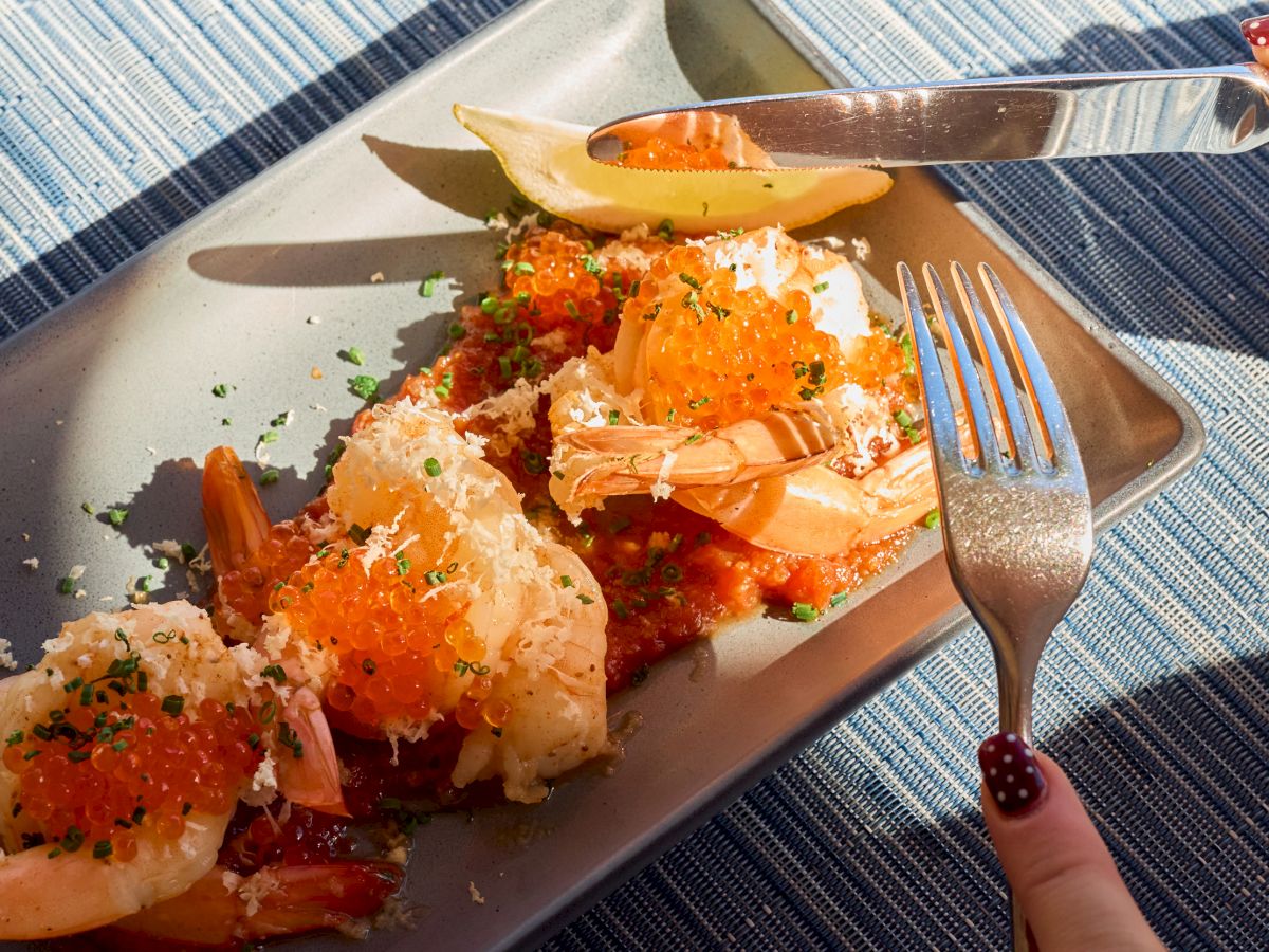 Delicious-looking sushi or crispy rolls topped with orange fish roe, sesame seeds, and shredded toppings on a rectangular plate, cutlery nearby.