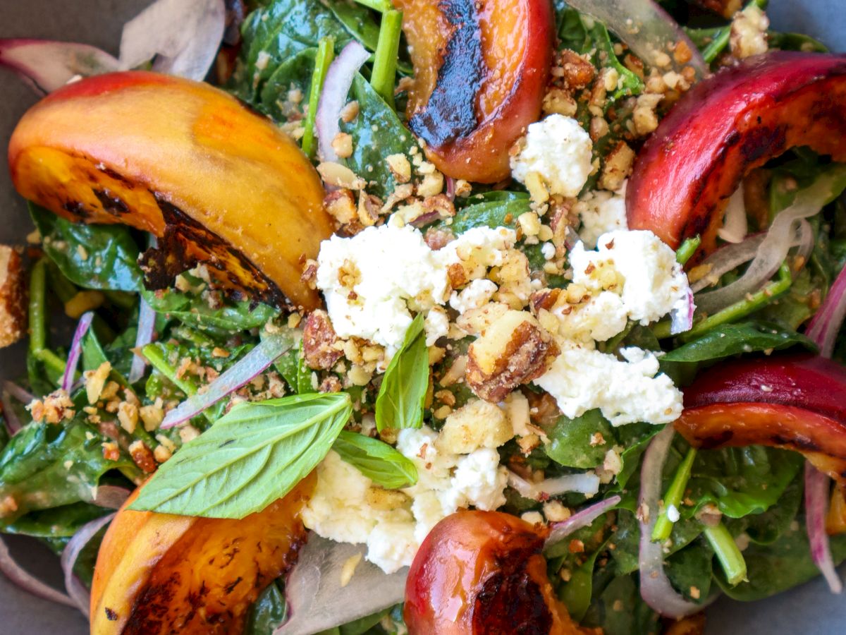 A salad with grilled peaches, greens, red onions, cheese, nuts, and basil in a gray bowl.