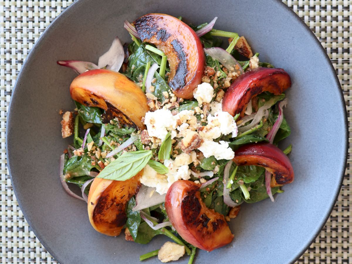 A salad with mixed greens, roasted peaches, and crumbled cheese, garnished with herbs and nuts in a gray bowl on a woven placemat.