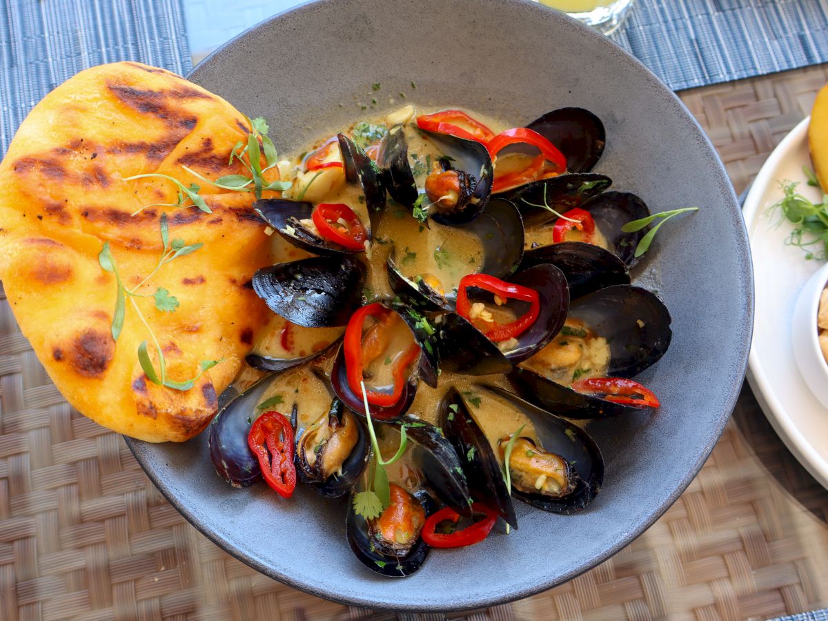 A bowl of mussels in a creamy sauce with sliced red peppers, garnished with herbs, served with a piece of grilled flatbread on the side.