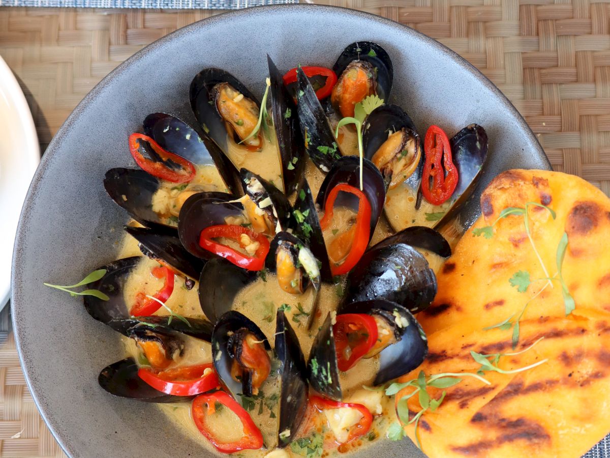 A bowl of mussels cooked in a creamy sauce with red peppers, garnished with herbs, and served with a piece of grilled bread.