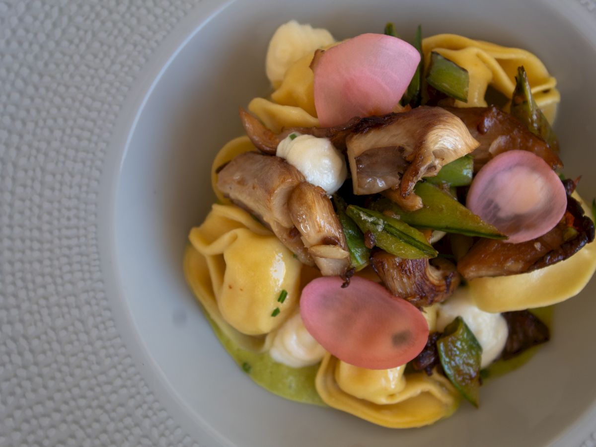 A bowl of tortellini topped with mixed vegetables, pink radish slices, and cheese or cream sauce, garnished with herbs.