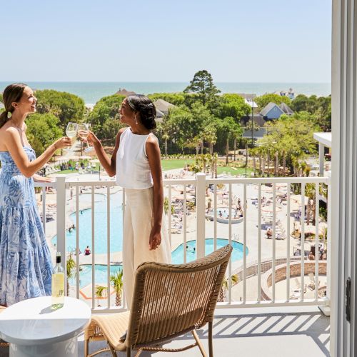 Two people are toasting on a balcony overlooking a pool and lush landscape by the sea.
