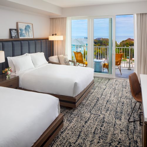 A modern hotel room with two beds, a desk, a lamp, and a view of the ocean from a balcony with chairs.