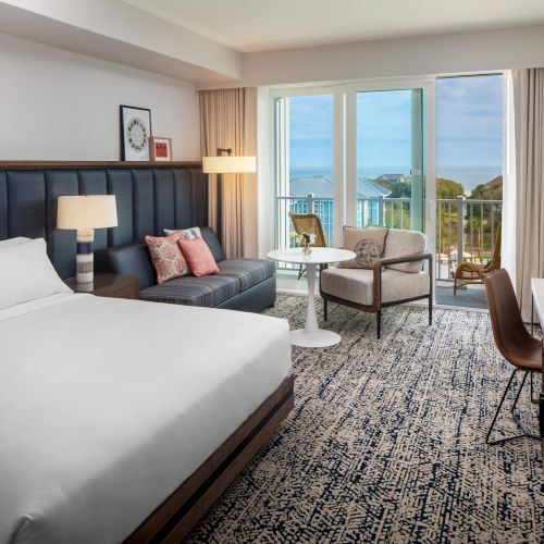 A modern hotel room with a bed, sofa, table, and chair, overlooking a balcony with a view of buildings and the sea in the distance.