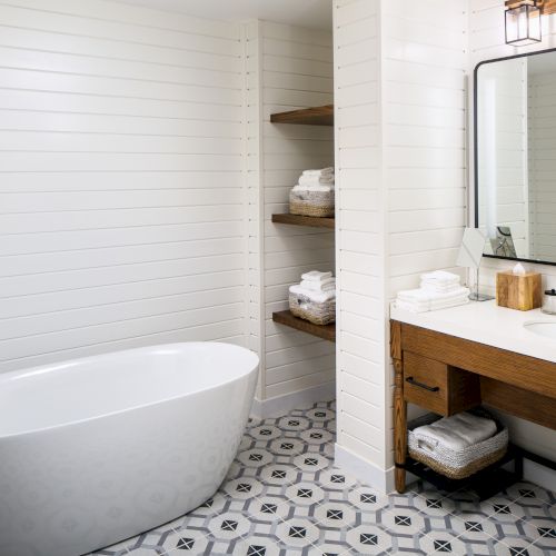 Modern bathroom with a freestanding white tub, wood vanity, large mirror, and decorative floor tiles; towels and amenities are neatly arranged.