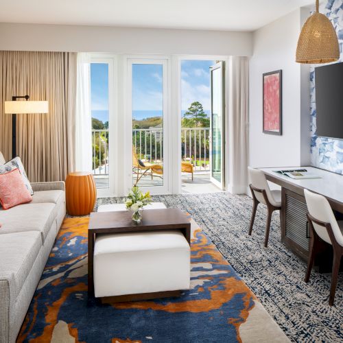 A modern living room with a sofa, TV, desk, and chairs opens to a balcony with a scenic view. Walls are blue-patterned with stylish decor.