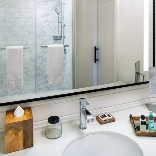A bathroom with a mirror, shower area, towels, faucet, tissue box, toiletries on a tray, and a small decorative item.