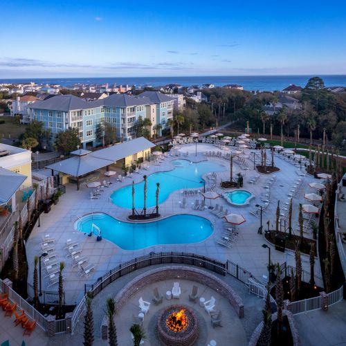 A resort with a large outdoor pool, lounge chairs, fire pit, buildings, and a view of the ocean in the background ends the scene.