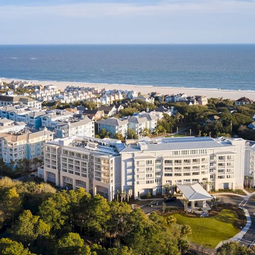 Aerial view of a beachfront hotel complex with multiple buildings, surrounded by trees and overlooking the ocean under a clear sky.