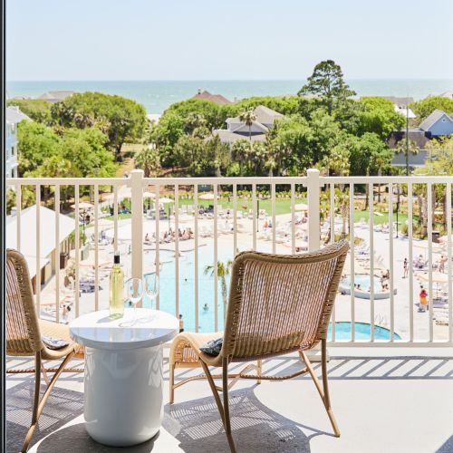 Two chairs and a small table on a balcony overlook a pool area and trees with the ocean in the distance.