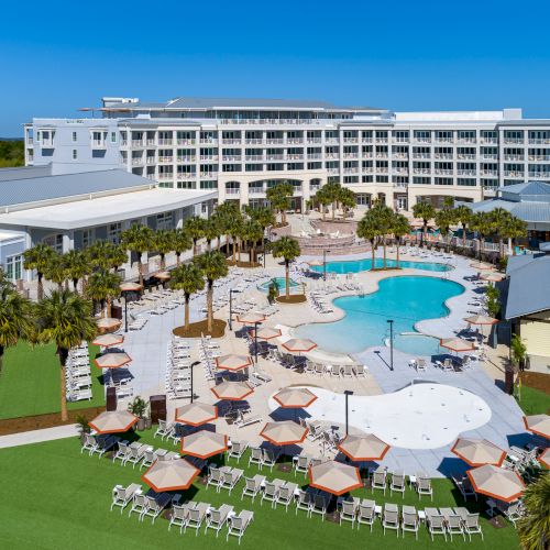 The image shows a resort with a curvy swimming pool, surrounded by lounge chairs, umbrellas, palm trees, and a large multi-story building.