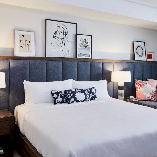A cozy bedroom with a large bed, blue headboard, side lamps, and artwork on the wall. There's also a bench with decorative pillows.