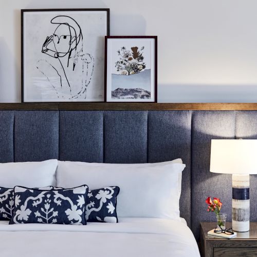 A neatly made bed with a dark upholstered headboard, decorative pillows, framed art, and a lamp on a bedside table holding flowers and glasses.